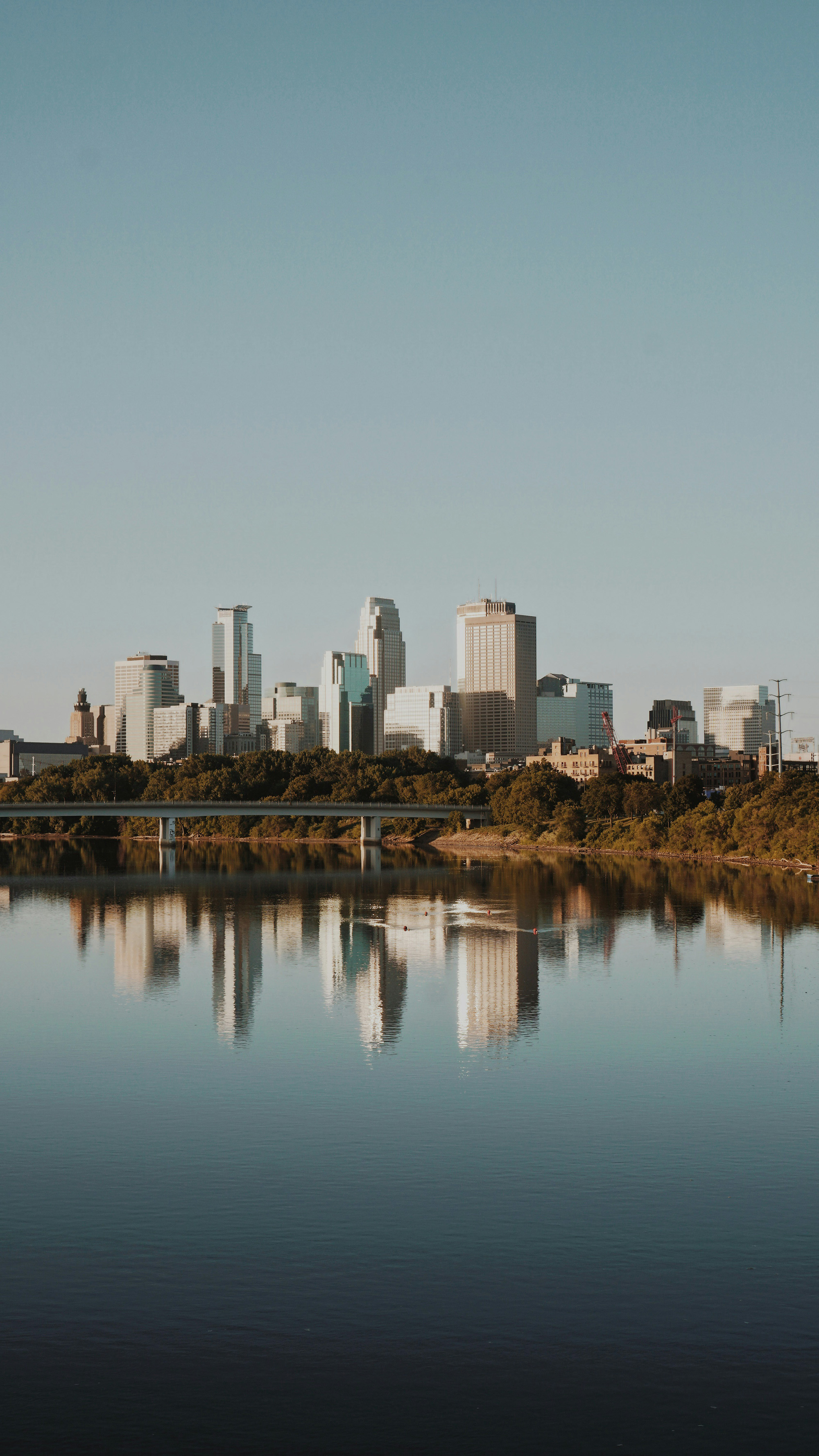 Skyline von Minneapolis mit Hochhäusern, Bäumen und Spiegelung im Mississippi bei klarem Himmel.