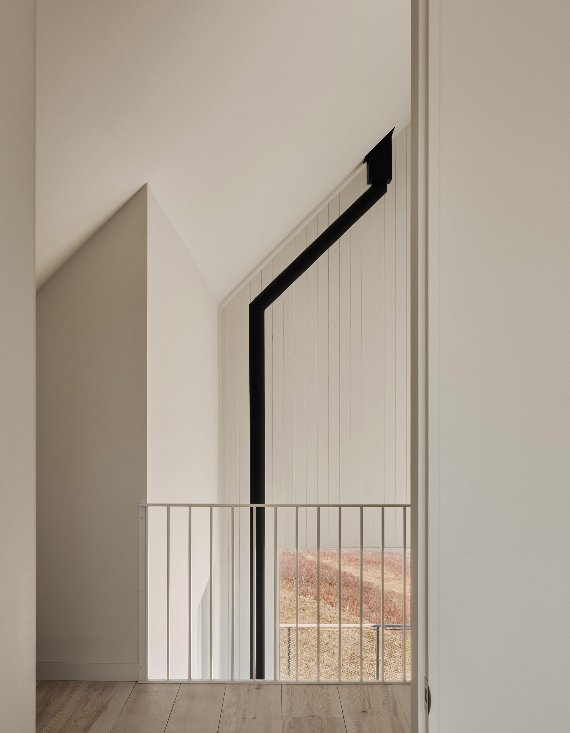 On the mezzanine, white-painted timber panelling, a slender balustrade and the black stove pipe meet in a restrained composition. The interior translates the simple gabled form into a clear spatial geometry.