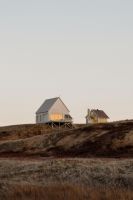 Graues Magdalen House auf Holzpfählen neben einem gelben Giebelhaus in der kargen Küstenlandschaft der Magdalen Islands.